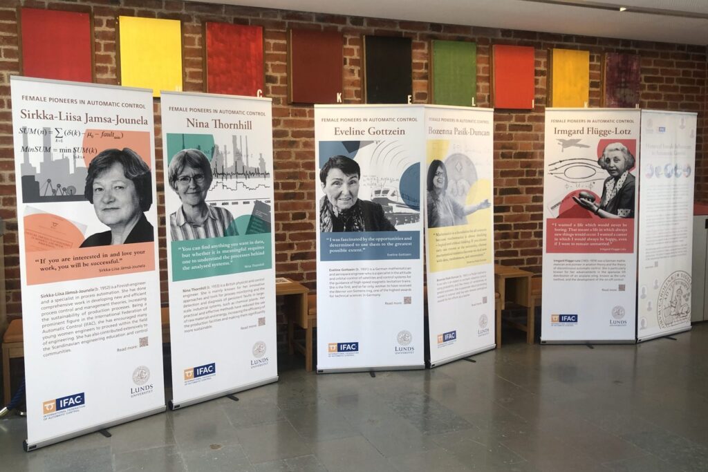 Five vertical banners display information and portraits of notable women in automatic control, standing against a brick wall with colourful rectangular panels above them. Each banner features a different woman’s name and achievements.