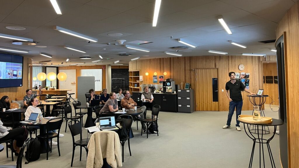 A man stands and presents in front of a group in a modern, well-lit classroom with wood-panelled walls. Attendees sit at tables, some with laptops open, listening attentively.