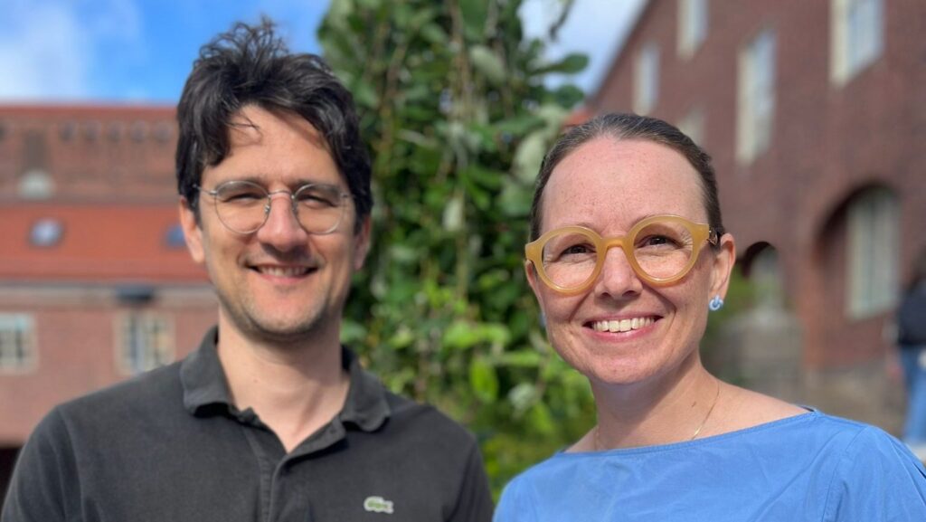 A man wearing glasses and a black polo shirt stands next to a smiling woman with round glasses and a blue top, outdoors with a leafy tree and red-brick buildings in the background.