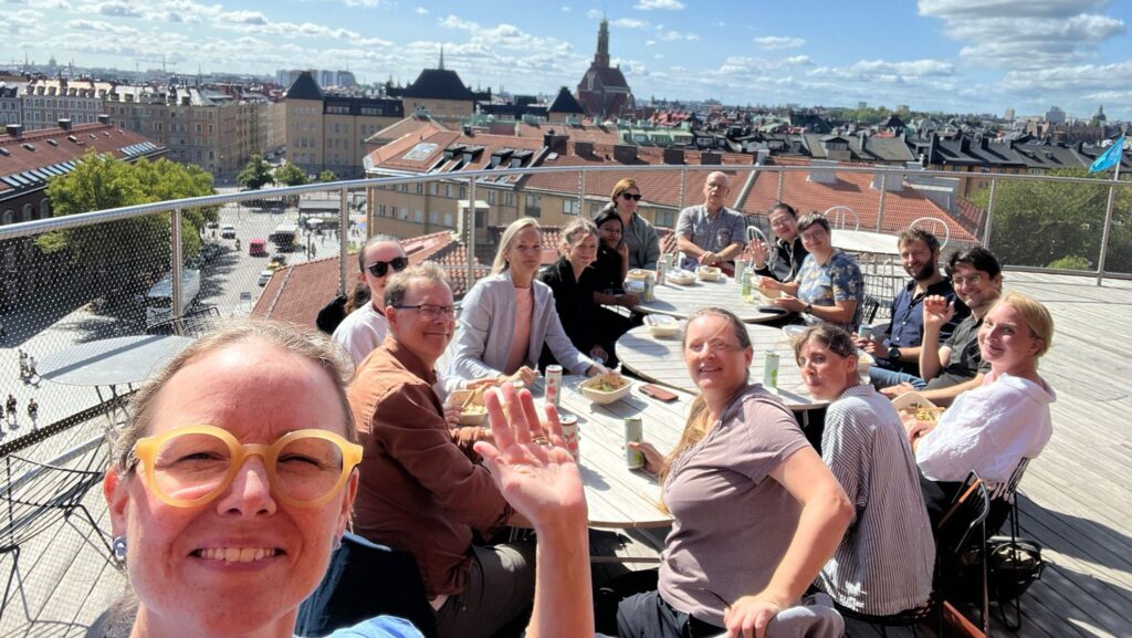A group of people sit around a table on a rooftop terrace, enjoying food and drinks under a sunny sky. One person in the foreground smiles and waves at the camera, with city buildings and a church spire in the background.