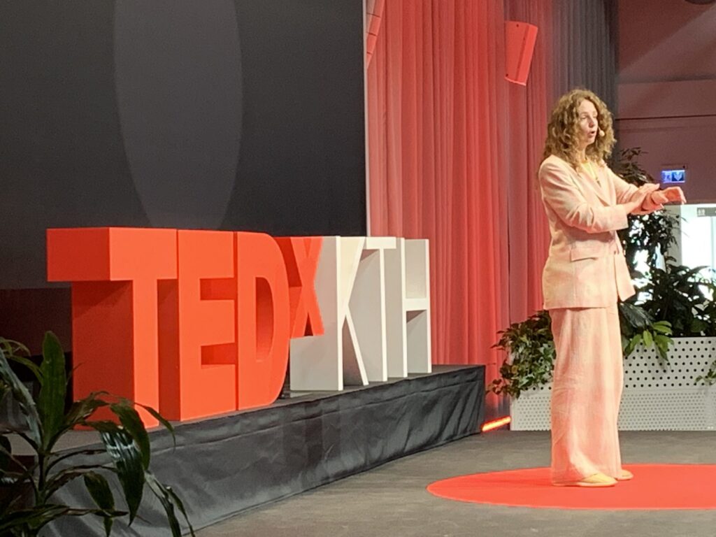 A speaker in a light pink suit stands on stage at a TEDxKTH event, gesturing with one hand. Large TEDxKTH letters are displayed beside her. The background and surroundings feature red drapes and green plants.