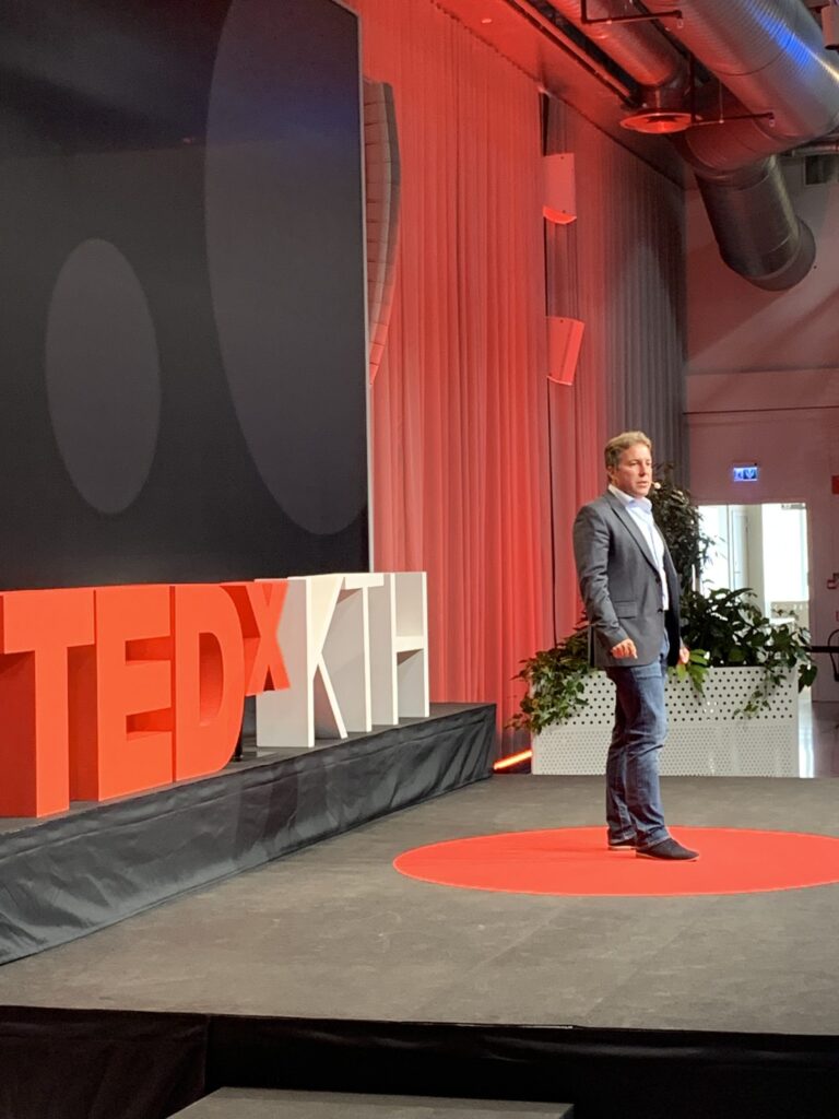 A speaker in a suit stands on a red circular carpet on stage at a TEDxKTH event, next to large TEDxKTH letters, with red curtains and industrial décor in the background.