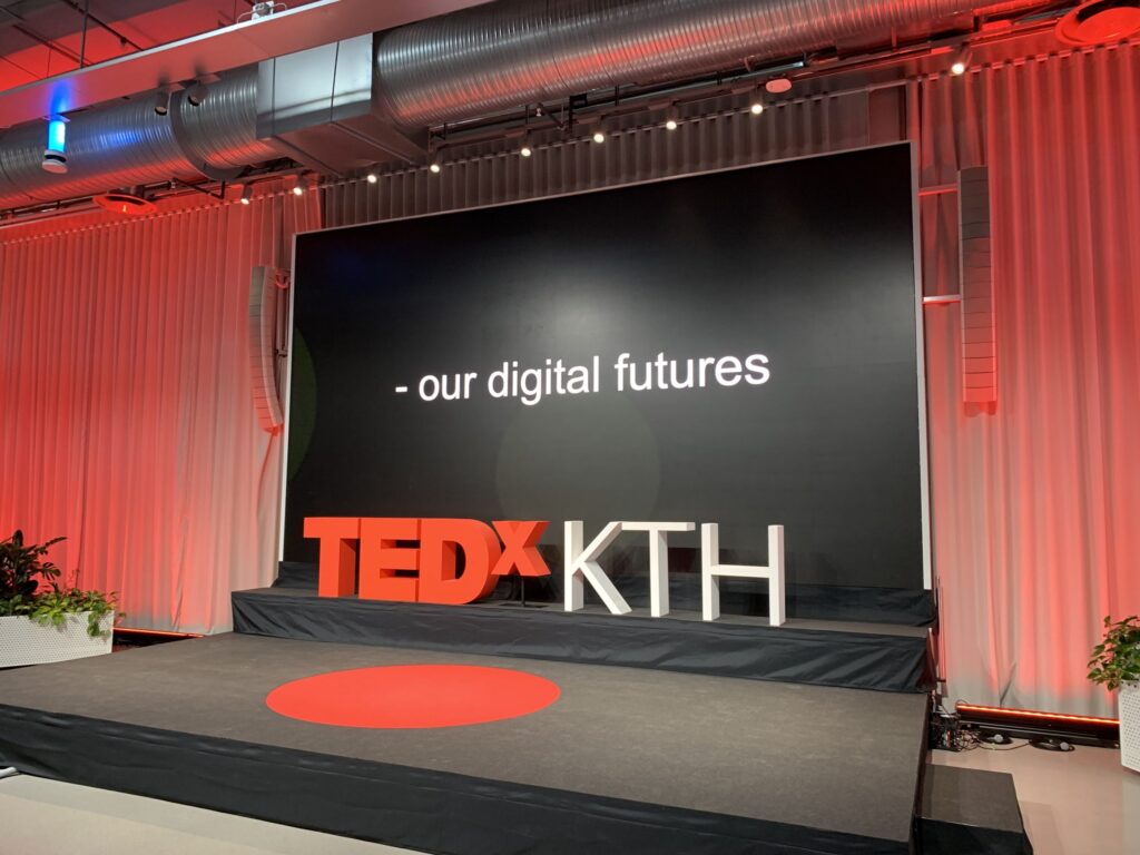 A TEDxKTH stage with a large screen displaying the text “- our digital futures.” The stage features bright red and white TEDxKTH letters and a red circle on the floor, with industrial lighting above.