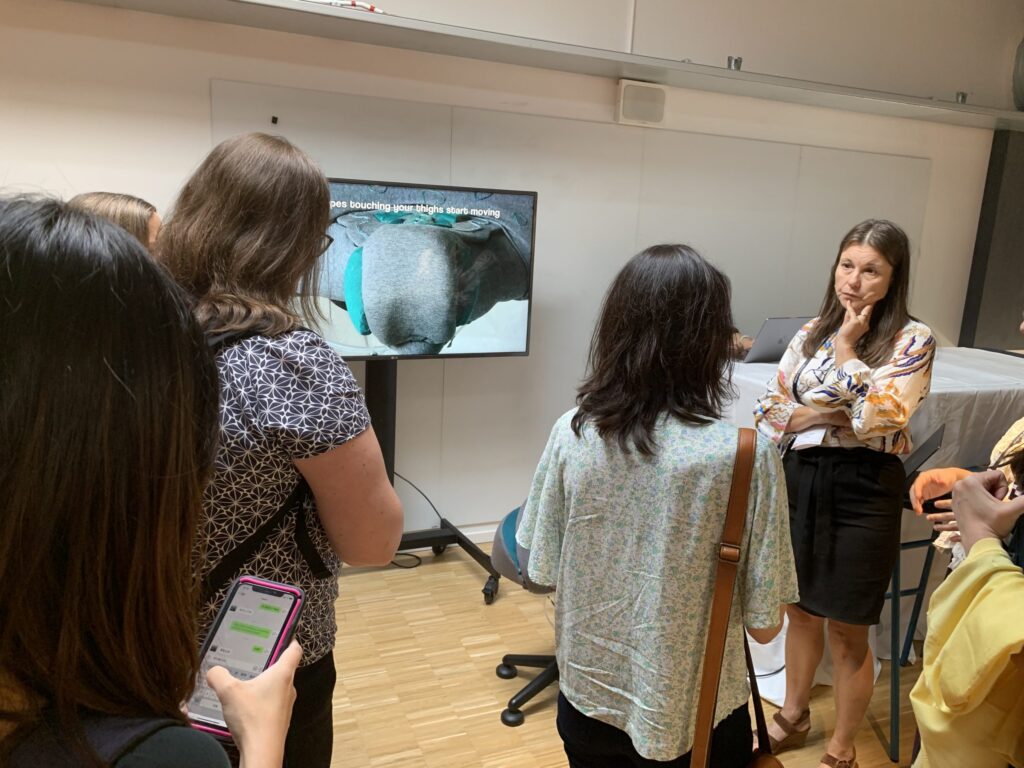 A group of women stand in a room, listening to a woman speak. Behind her is a screen displaying an image of an elephant and text. One woman in the foreground holds a mobile. The setting appears to be a classroom or workshop.