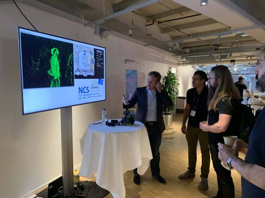 Three people stand around a small round table while one person gestures at a large screen displaying green 3D imagery and graphics about NCS in a modern, well-lit office setting.