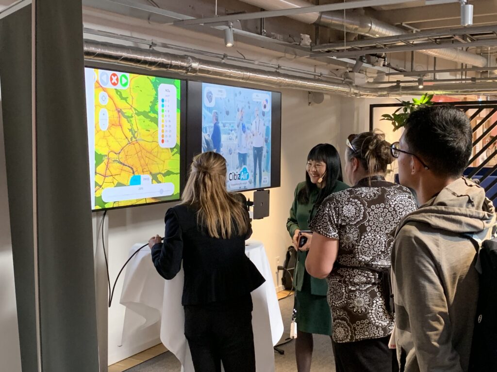 Four people stand in front of two large screens; one shows a colourful map, and the other displays a presentation. One woman points at a screen while the others watch attentively in a modern indoor setting.