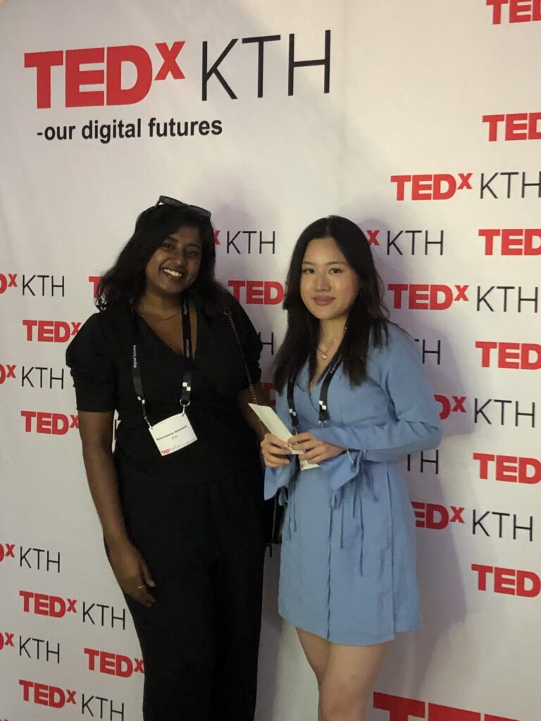 Two women stand and smile in front of a TEDxKTH event backdrop. One wears a black outfit and conference lanyard, the other wears a light blue dress and holds a small book.