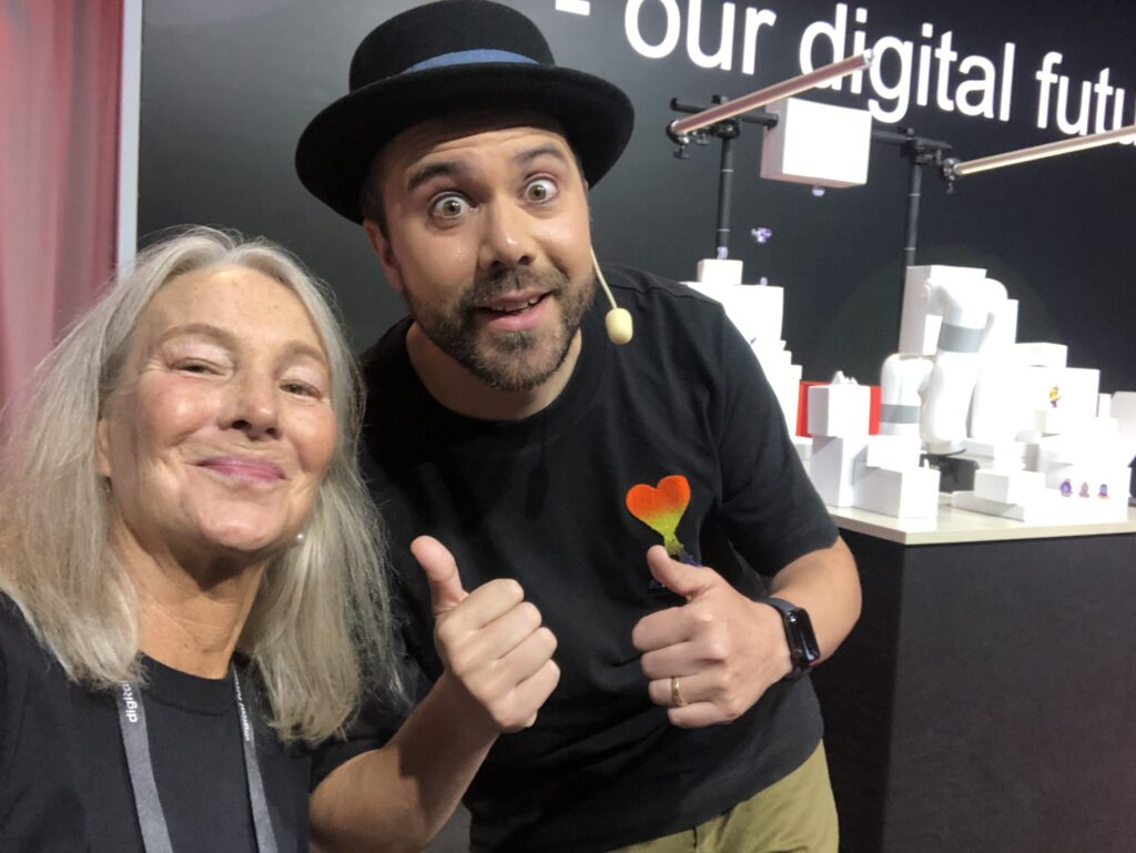 A woman with long grey hair and a man with a beard and black hat smile and give thumbs up in front of a display with white geometric objects and a sign that reads “our digital futu”.
