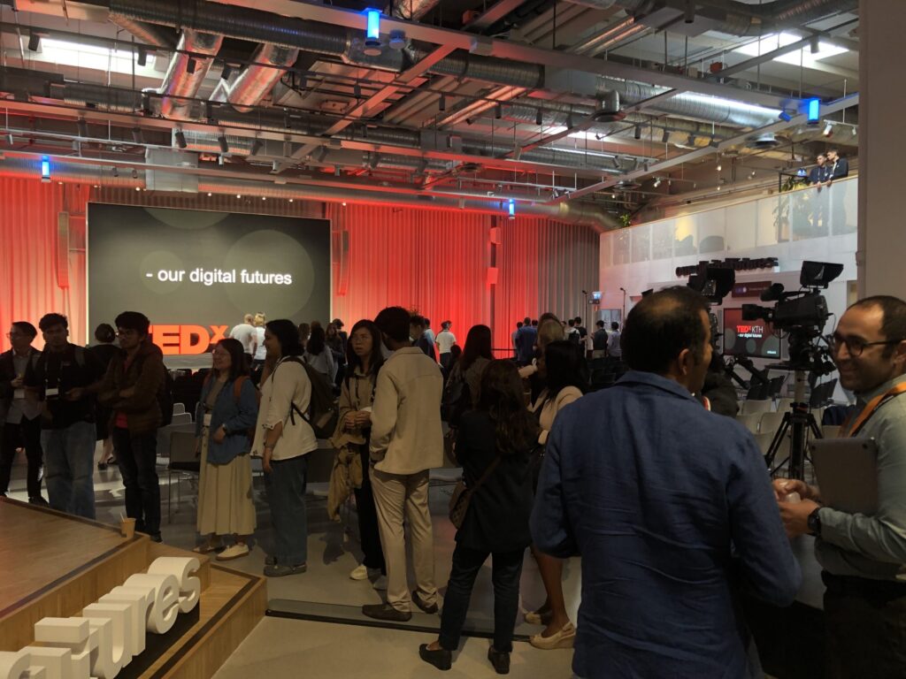 A group of people gather and network in a modern event space with cameras and lighting equipment. A TEDx stage in the background displays the words our digital futures.