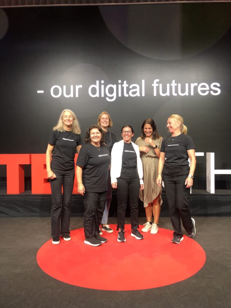 Six women stand smiling on a TEDx stage, five wearing matching black t-shirts and trousers, one in a beige dress. The backdrop displays the words our digital futures in white text on a black background.