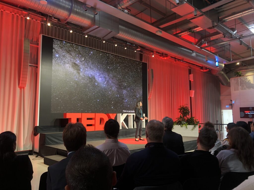 A speaker stands on stage at a TEDx event, with a large screen behind showing a starry night sky. Audience members are seated, and the room is lit with red and white lighting.