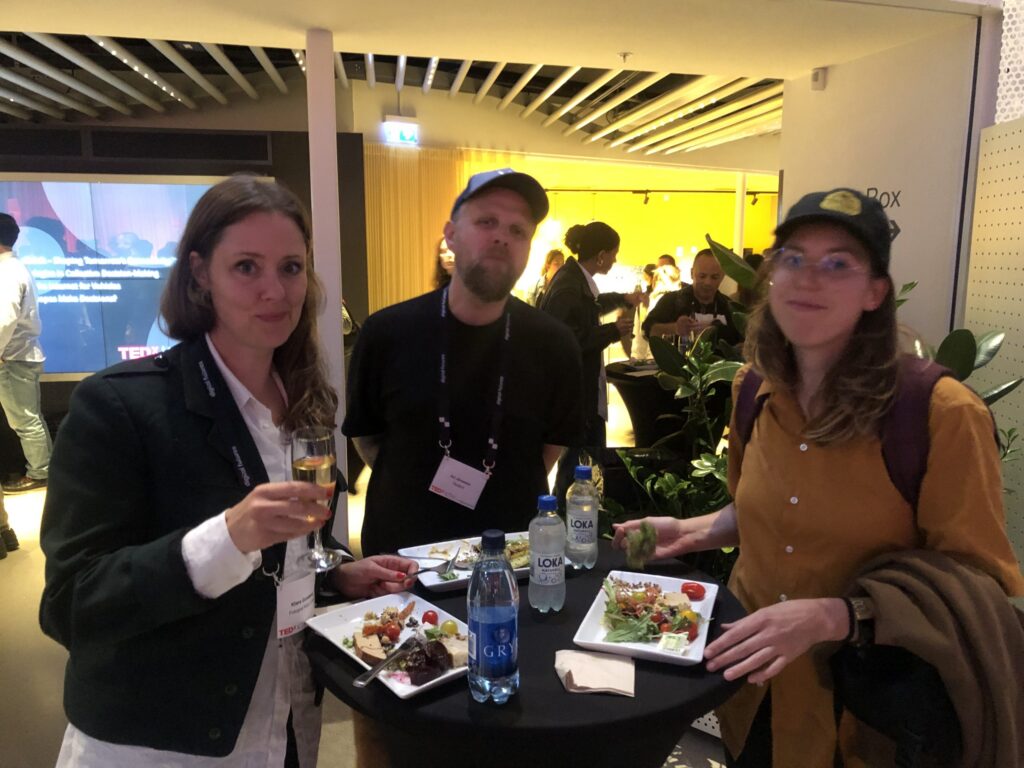 Three people stand around a high-top table with plates of food and water bottles at a social event. Two women and one man look at the camera, while other attendees mingle in the background.