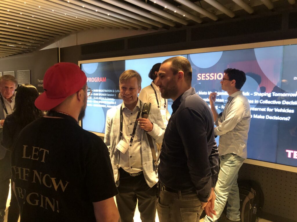 A group of people stand and talk in front of a large screen displaying event programme information. One man wears a red hat and black shirt with LET THE NOW BEGIN! printed on the back.