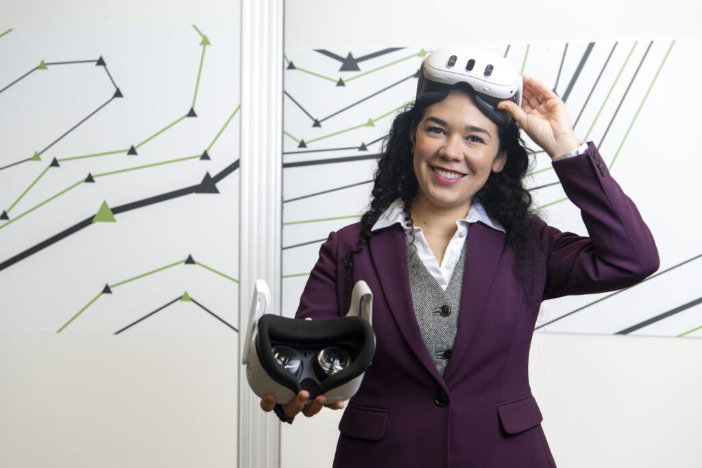A woman in a maroon blazer smiles while holding a virtual reality headset in one hand and placing another on her head. She stands in front of a white wall with abstract black and green arrow designs.