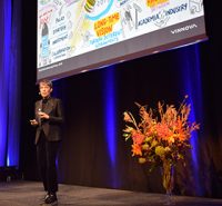 A person in dark clothing speaks on stage next to a tall bunch of orange flowers, with a large illustrated presentation projected behind them. Blue stage lights illuminate the background.