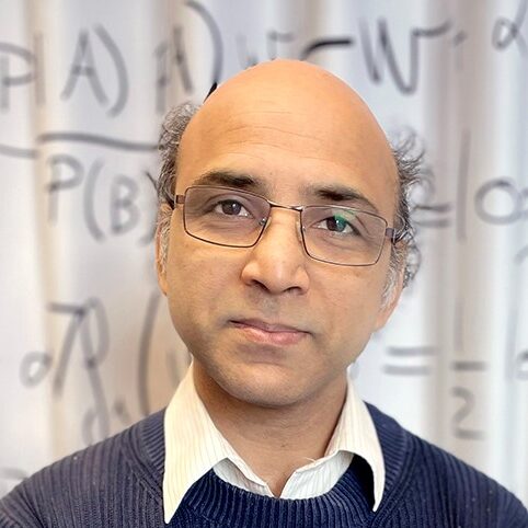 A man with glasses, a bald head, and wearing a blue jumper over a collared shirt stands in front of a whiteboard covered with mathematical equations.