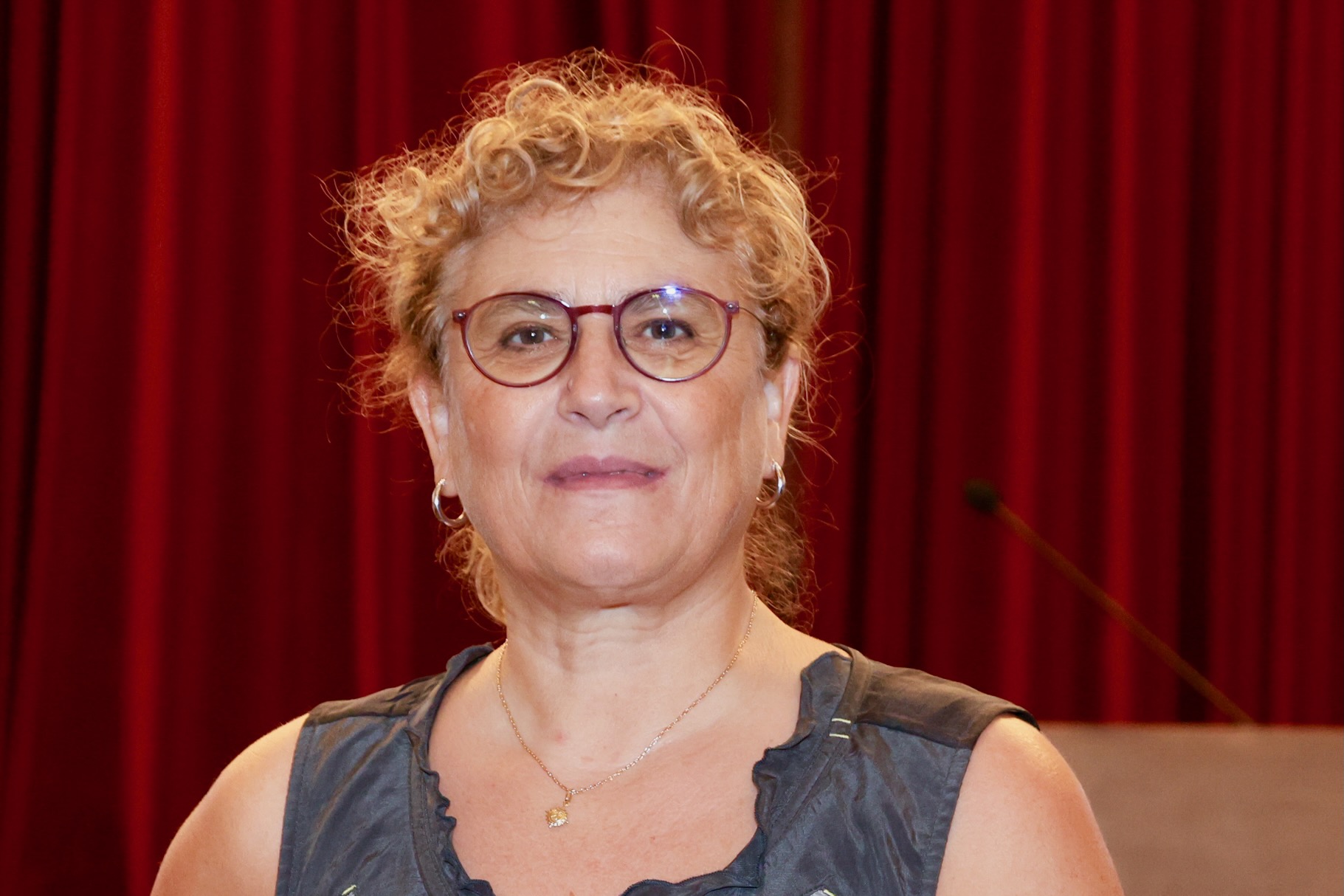 A woman with curly blonde hair, glasses, and a sleeveless grey top is standing in front of red curtains, looking at the camera and smiling slightly.