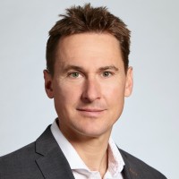 A man with short brown hair wearing a dark suit jacket and a white collared shirt, looking at the camera with a neutral expression against a light grey background.