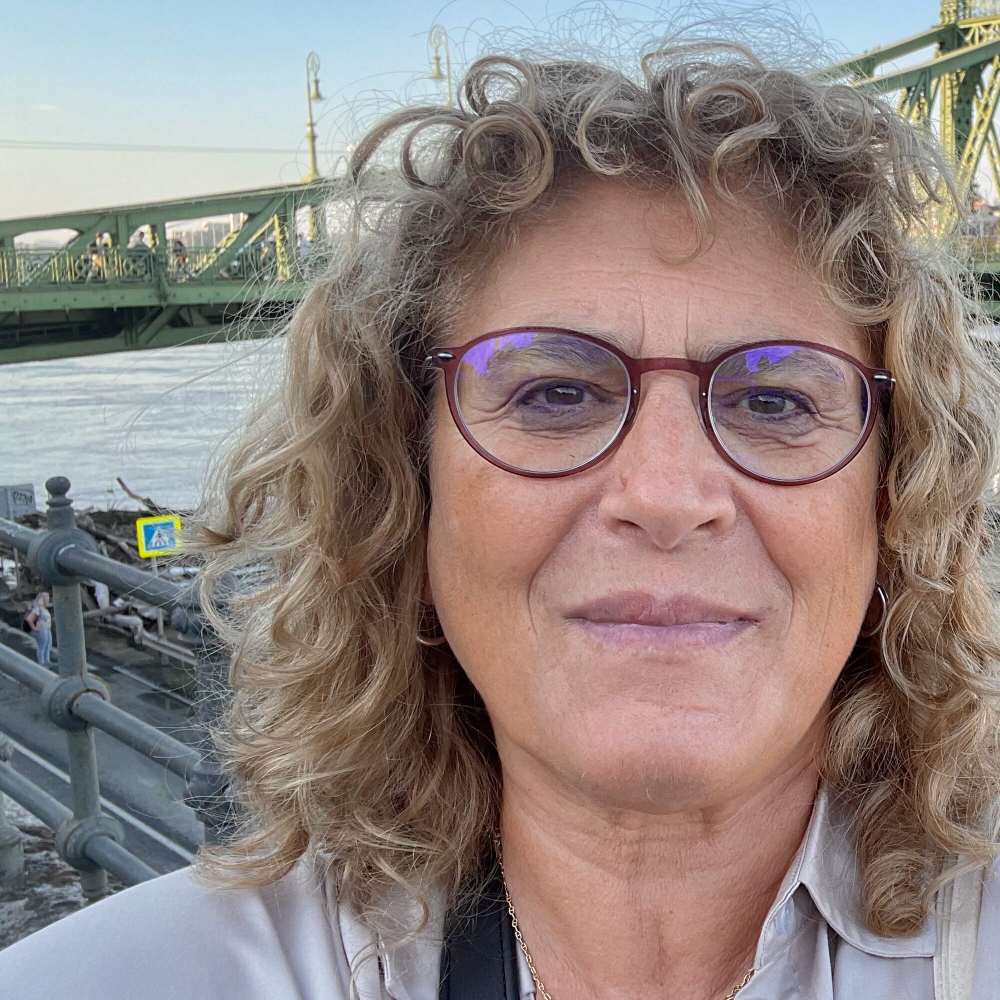 A woman with curly blonde hair and glasses smiles at the camera, standing outdoors near a river with a green metal bridge in the background.