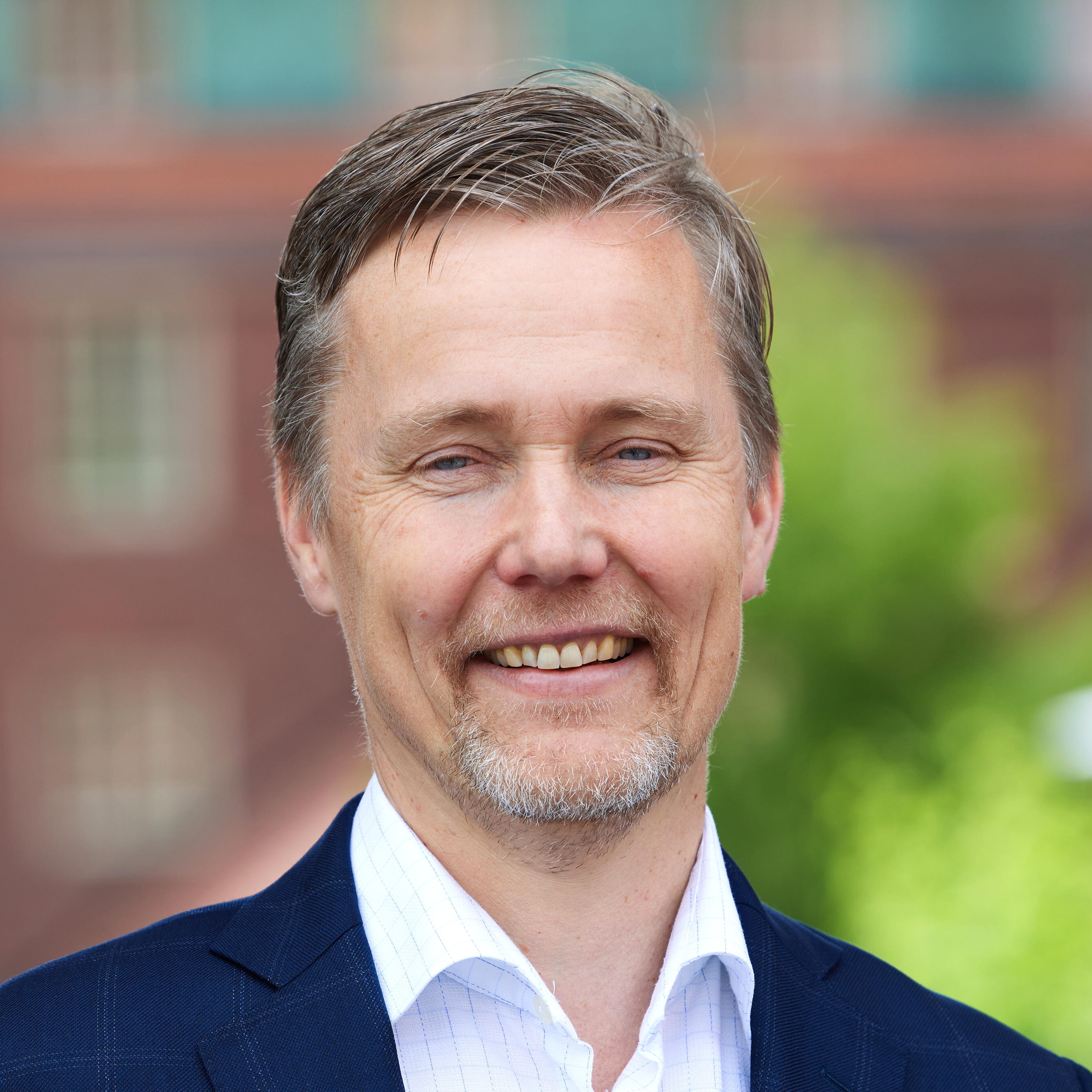 A middle-aged man with short grey hair and a trimmed beard wears a blue suit and white shirt, smiling outdoors with a blurred brick building and greenery in the background.