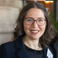 A woman with wavy brown hair and glasses smiles at the camera. She is wearing a dark blazer over a beaded top and a name badge. The background is slightly blurred, suggesting an indoor setting.