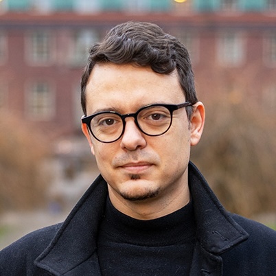 A man with short brown hair and glasses, wearing a black coat and polo neck, stands outdoors with a blurred building and trees in the background.