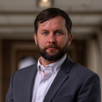 A man with a beard and short brown hair, wearing a grey suit jacket and white dress shirt, stands indoors with a blurred background.