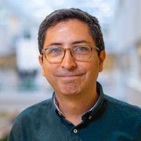 A man with short dark hair and glasses, wearing a dark green button-up shirt, smiles softly at the camera. The background is softly blurred, showing an indoor setting with natural light.