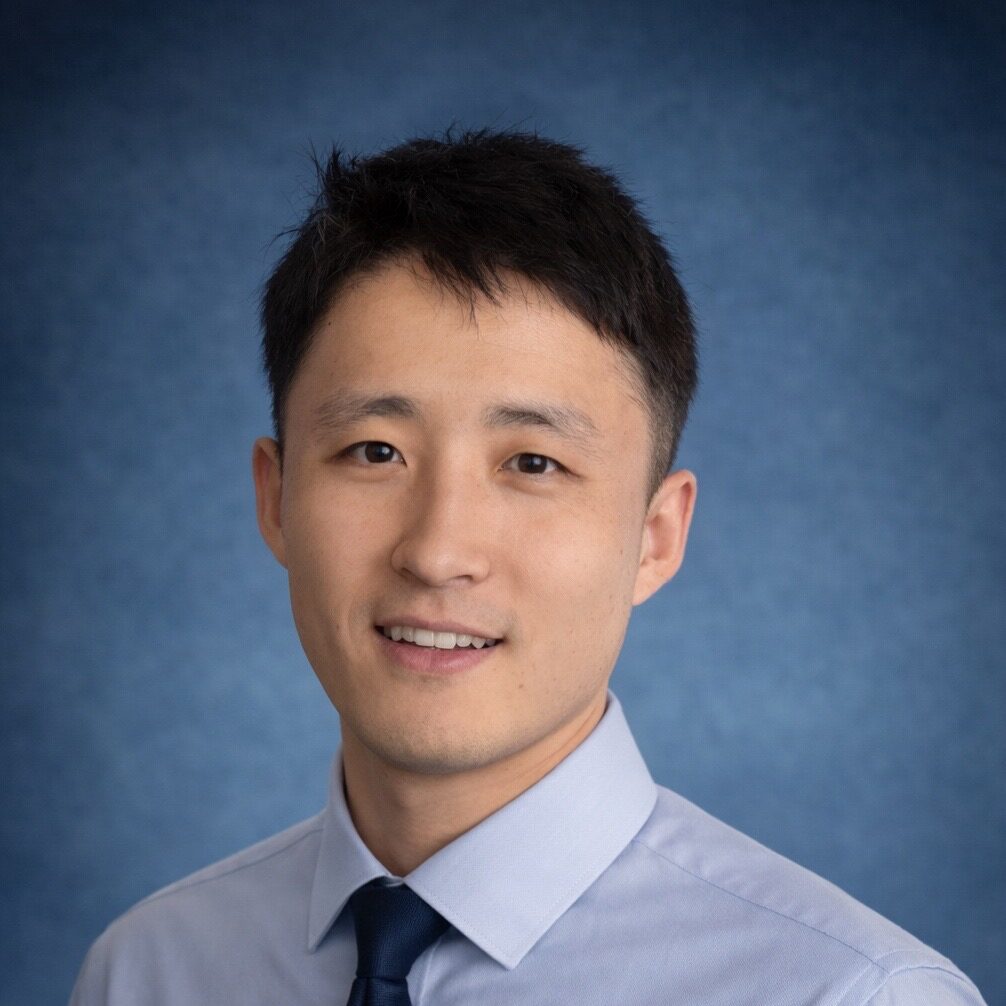 A man with short black hair wearing a light blue dress shirt and a dark blue tie smiles in front of a blue studio background.