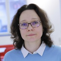 A woman with shoulder-length brown hair and glasses, wearing a light blue jumper over a white collared shirt, stands indoors with a blurred background.