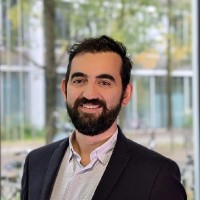 A man with dark hair and a beard, wearing a suit jacket and light shirt, smiles in front of a window with trees and modern buildings visible outside.