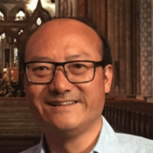 A man with glasses and a light blue shirt smiles whilst standing inside a grand, ornate cathedral with wooden pews and tall stone columns in the background.