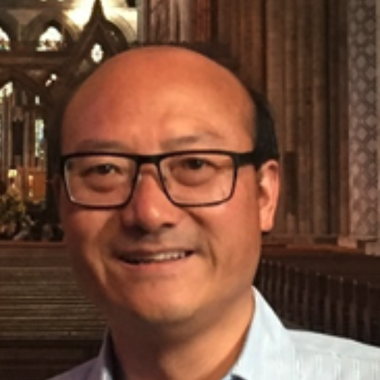 A man with glasses and a light blue shirt smiles whilst standing inside a grand, ornate cathedral with wooden pews and tall stone columns in the background.