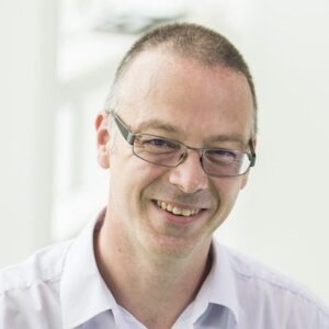 A middle-aged man with short hair and glasses smiles at the camera. He is wearing a light purple collared shirt and is pictured in a bright, softly lit indoor setting.