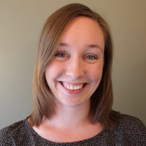 A woman with straight, shoulder-length light brown hair smiles at the camera. She is wearing a black and white patterned top and is standing in front of a plain light-coloured background.