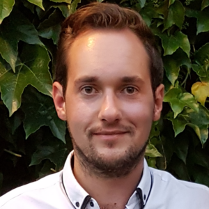 A man with short brown hair and a trimmed beard, wearing a light-coloured shirt, stands in front of a background of green leaves, smiling slightly at the camera.