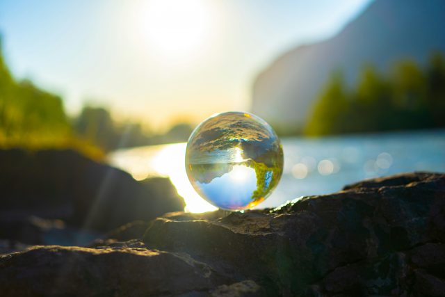 A glass sphere rests on a rock by a river, reflecting an inverted image of the sunlit landscape, with blurred trees, water, and mountains in the background during a bright day.