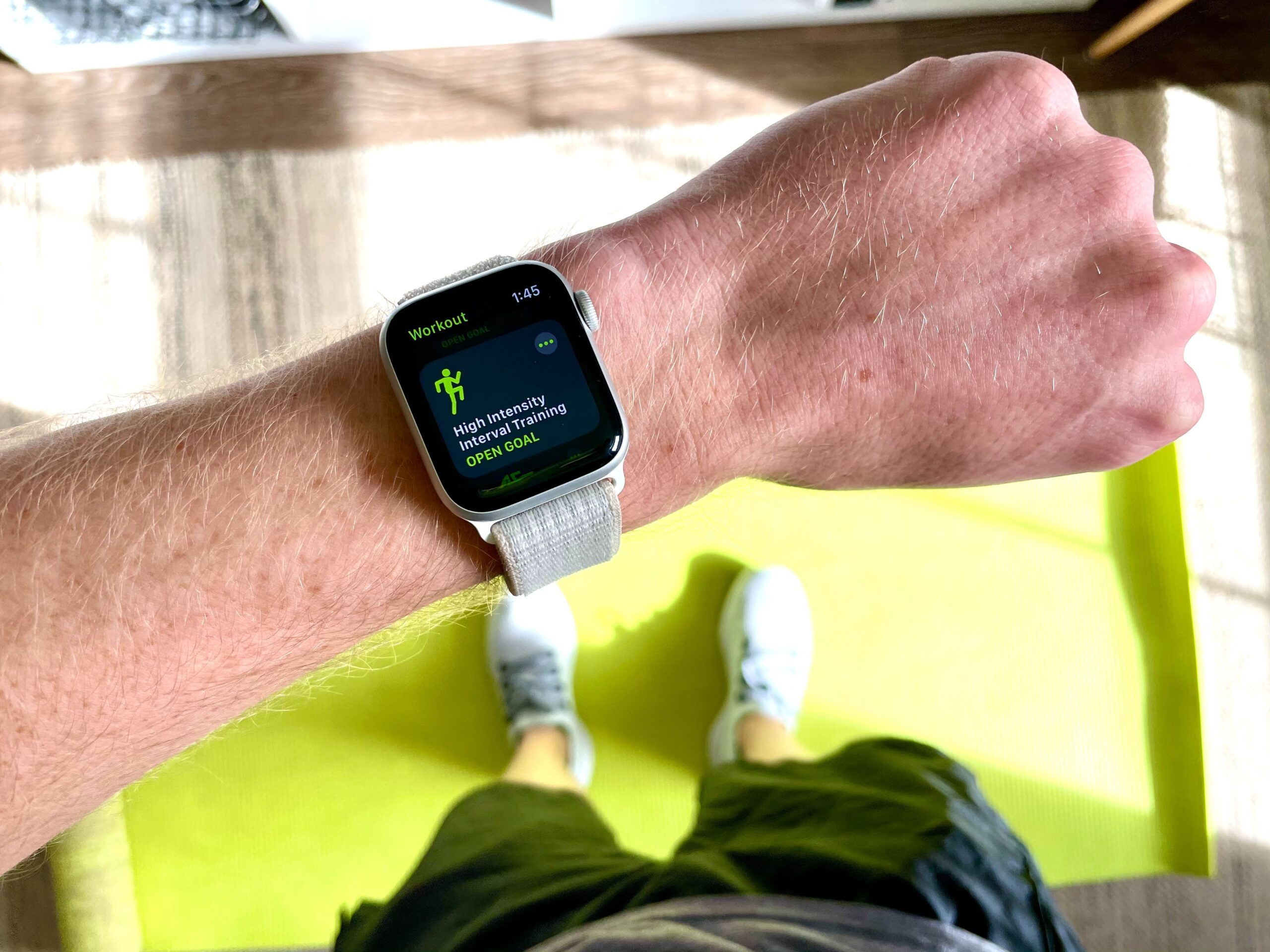 A person wearing a smartwatch displaying a workout summary stands on a bright green exercise mat indoors, dressed in shorts and trainers.