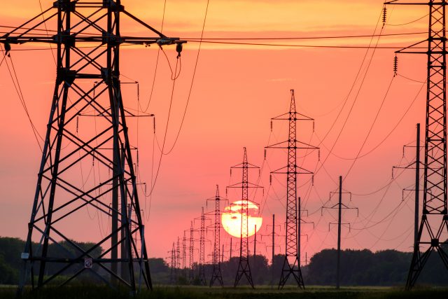 Rows of electrical power lines and pylons stretch into the distance across a field, silhouetted against a vivid orange and pink sunset, with the sun low on the horizon above tree lines.