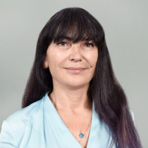 A woman with long dark hair and fringe, wearing a light blue top and a pendant necklace, smiles softly in front of a plain light grey background.