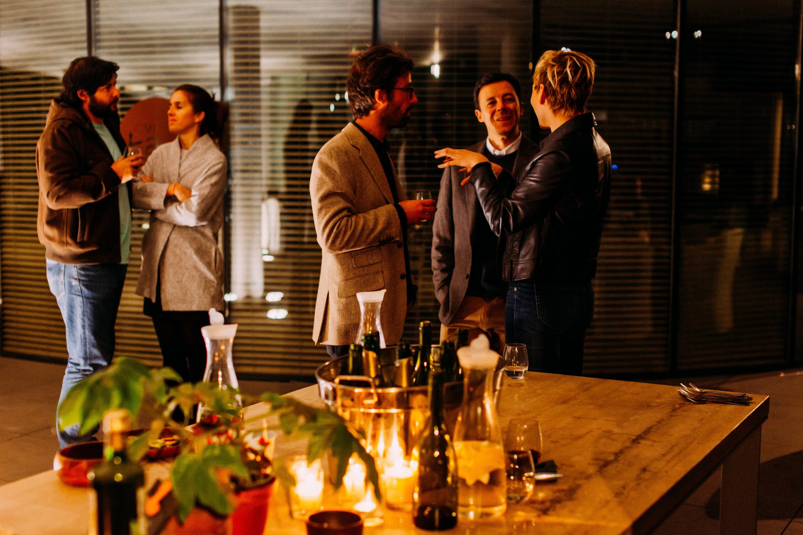 A group of six people are socialising at a warmly lit indoor gathering, talking in small groups near a table set with candles, bottles, and drinks.