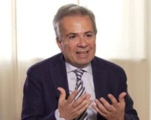 A man in a suit and striped tie gestures with both hands while speaking, seated in front of a softly lit curtain background.