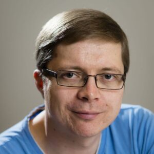 A man with short brown hair and glasses smiles slightly at the camera. He is wearing a light blue shirt and is pictured against a plain, neutral background.