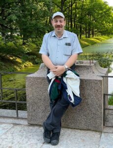 A man in a light-coloured cap and striped short-sleeve shirt stands smiling by a stone railing in a green park with trees and a pond in the background. He holds a jacket in his arms.