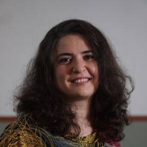 A woman with long, wavy dark hair smiles at the camera. She is wearing a colourful top with a geometric pattern, and the background is plain and softly lit.