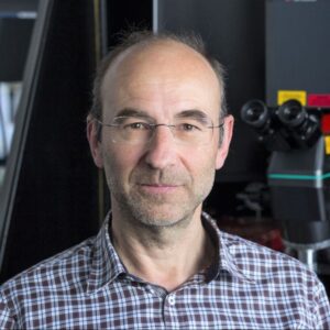 A middle-aged man with glasses and a chequered shirt stands indoors, with scientific equipment and bookshelves visible in the background.