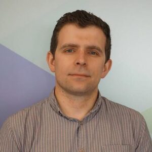 A man with short brown hair is wearing a checked button-up shirt and looking at the camera. The background features geometric shapes in pastel blue, green, and purple tones.