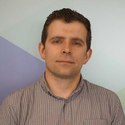 A man with short brown hair is wearing a checked button-up shirt and looking at the camera. The background features geometric shapes in pastel blue, green, and purple tones.