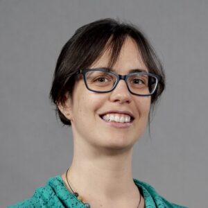A woman with straight brown hair, wearing glasses and a teal top, smiles at the camera against a plain grey background.