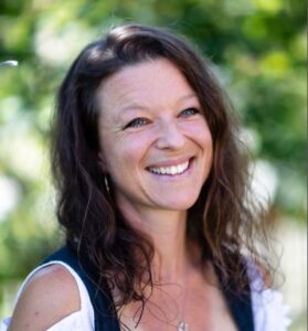 A woman with long wavy brown hair smiles outdoors, wearing a dark top over a white shirt, with a blurred green background of foliage.
